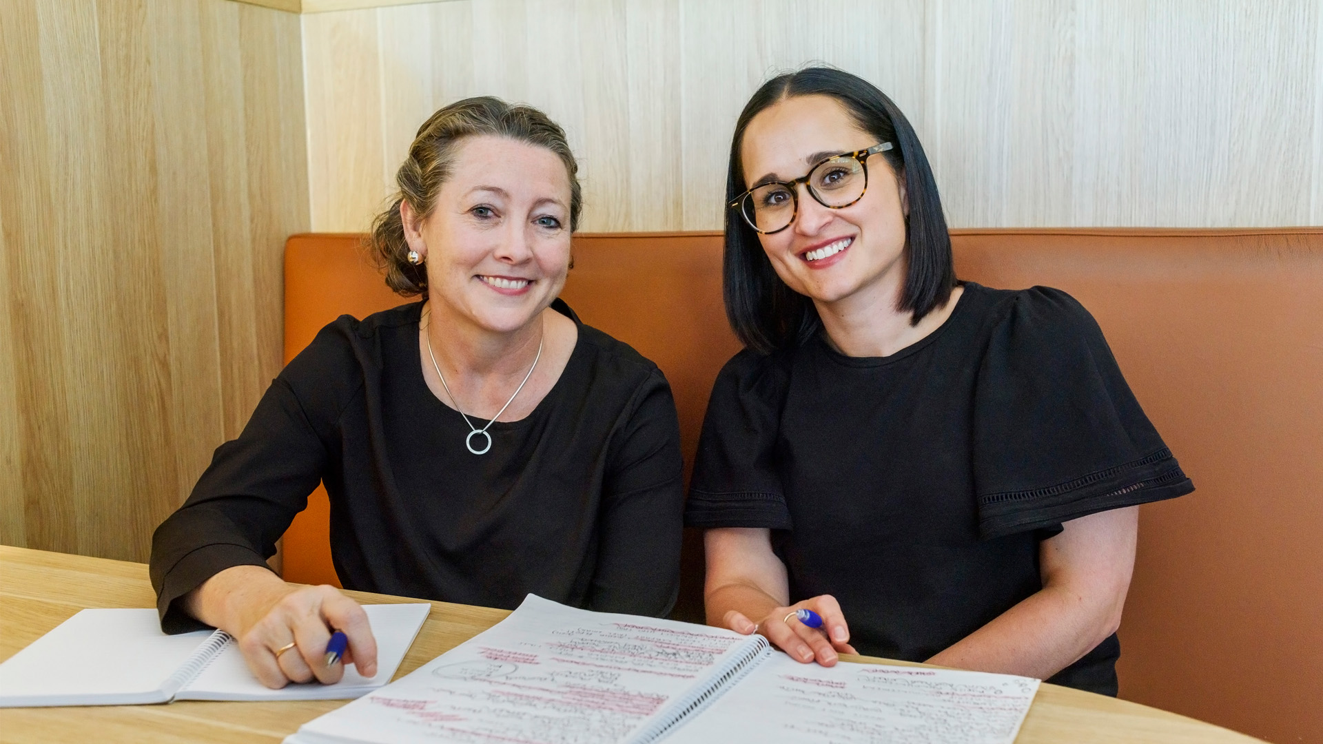 Two women seated at a café table, engaged in conversation with a notebook open in front of them.