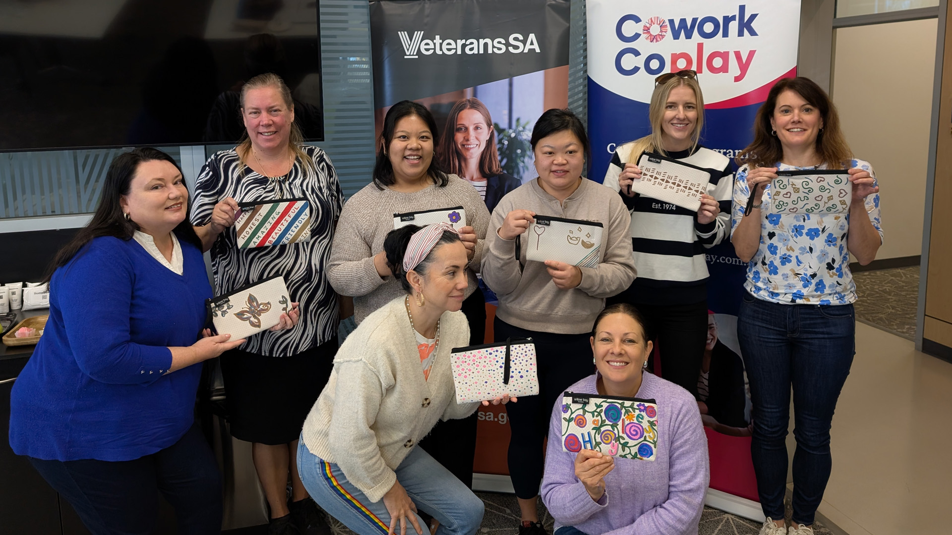 Group of eight women posting indoors, smiling and holding a handmade craft item, standing and kneeling in front of display banners in a community setting.