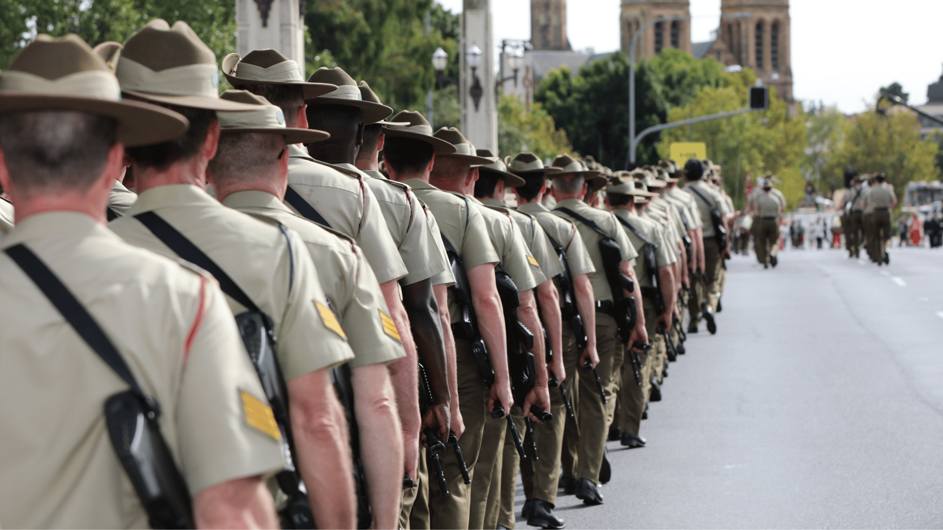 A long line of uniformed Australian Army personnel marching in formation along a city street during a commemorative parade, viewed from behind with buildings and spectators in the distance.