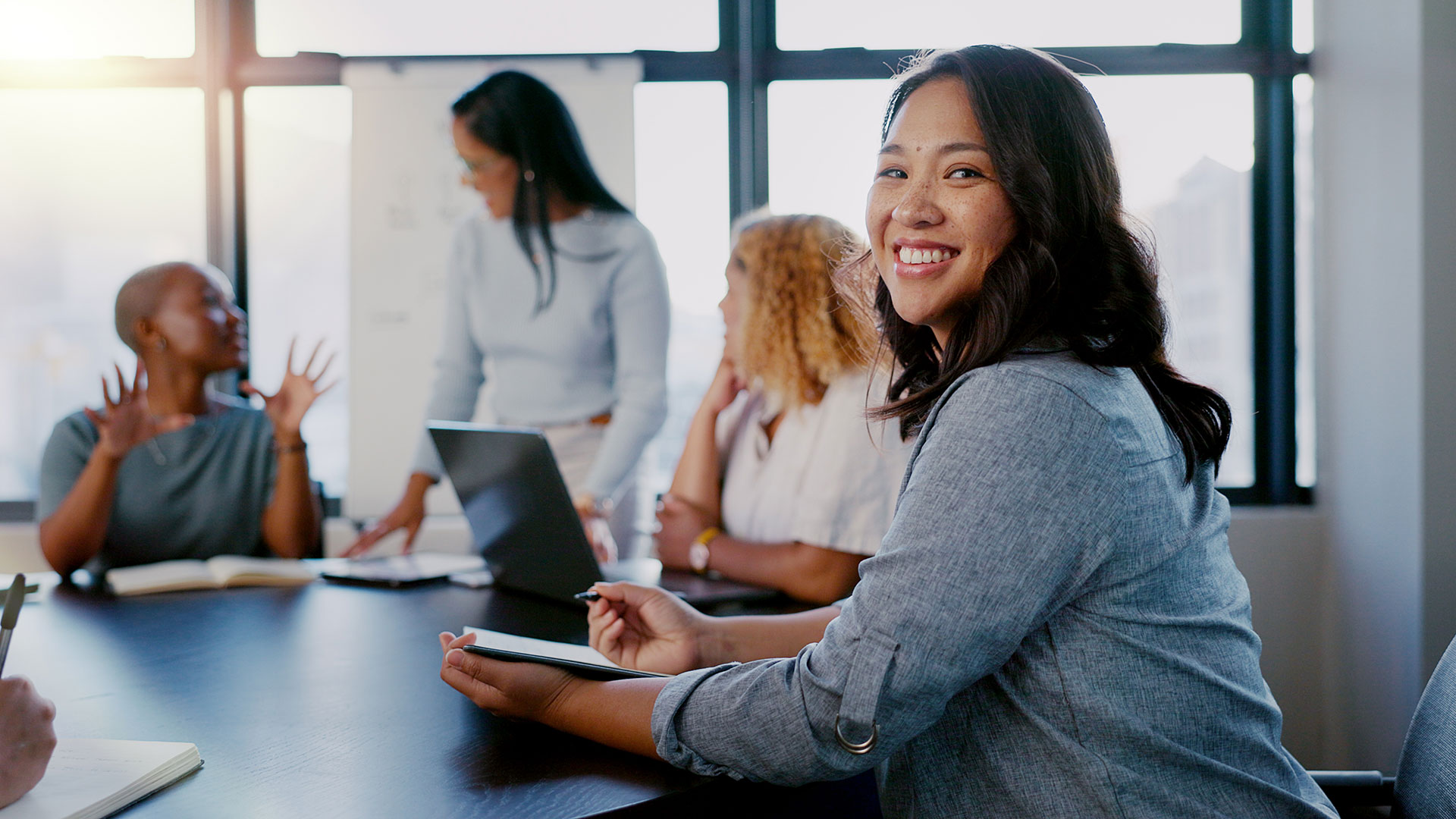 Australian-Asian woman sitting at a desk, smiling at the camera while people are in conversation behind her