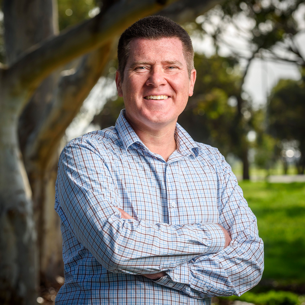 Veterans SA Tertiary Scholarship recipient and RAAF veteran, Ewan Person in a light checkered button-up shirt standing outdoors with arms crossed beside large tree trunks