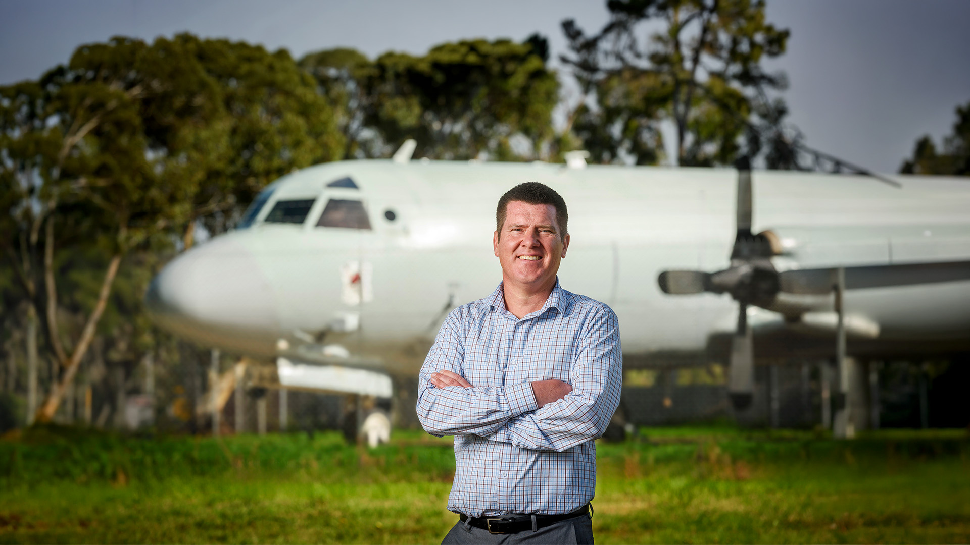 Person standing outdoors with arms crossed in front of a parked aircraft.