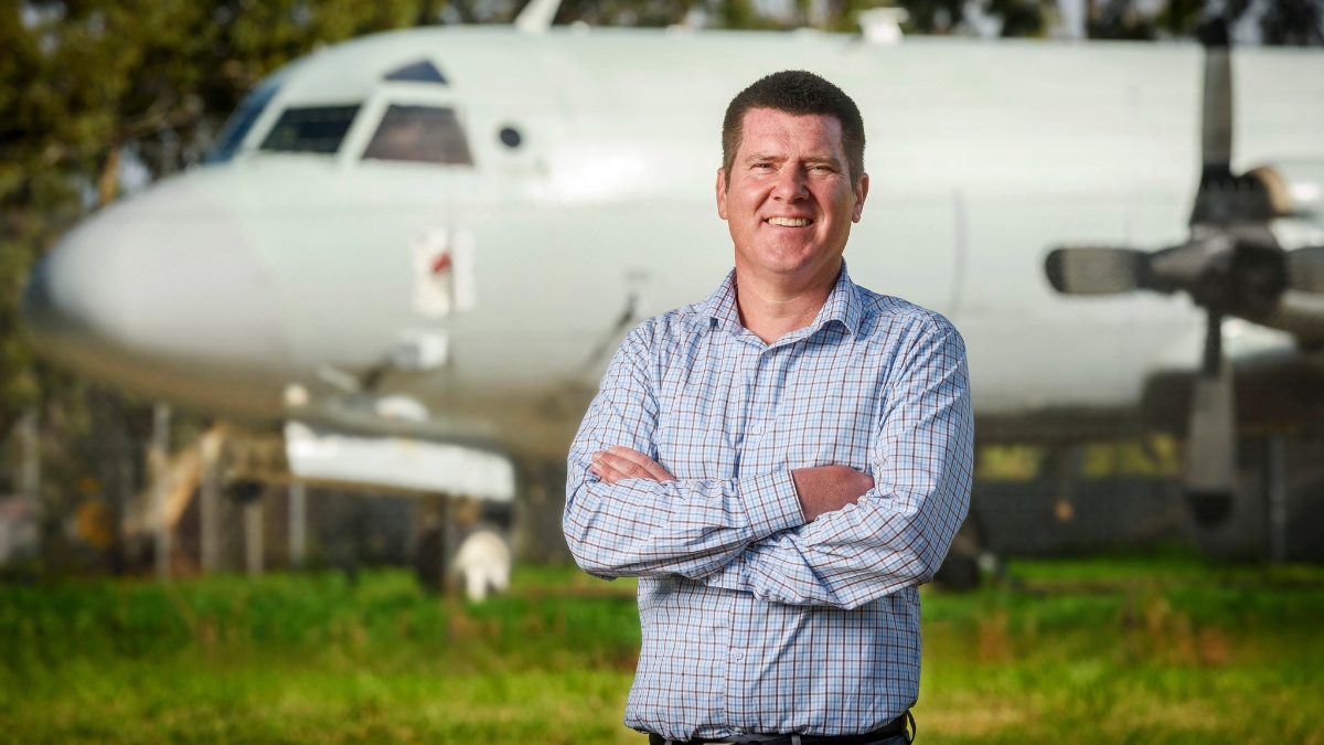 Person standing outdoors with arms crossed in front of a parked aircraft.